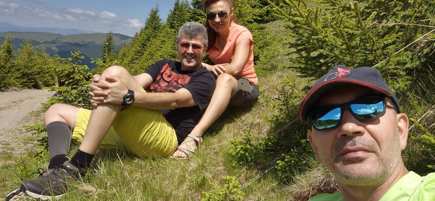 Alex in the foreground with Radu and Violeta resting on a grassy slope among young fir trees, mountains behind