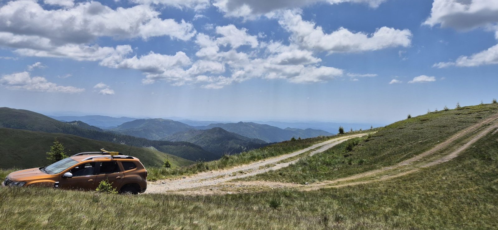 Our car beside a grassy ridge road with forested mountains and summer clouds beyond